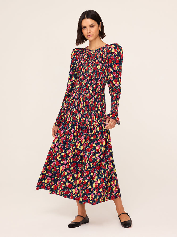 A woman stands against a plain background, wearing the Wren Painted Floral Print Shirred Dress by KITRI. The midi dress features voluminous sleeves and a vibrant pattern with red, pink, yellow, and green flowers. She pairs it with black flat shoes, and her short dark hair is styled in a bob.