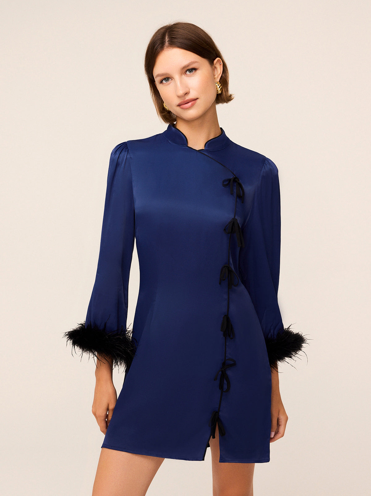 A woman stands against a plain background, elegantly dressed in the KITRI Naomi Navy Satin Feather Mini Dress. The dress features sophisticated feathered cuffs and a cheongsam-inspired collar with black ribbon closures that enhance its allure. Her short brown hair beautifully complements her gold earrings.