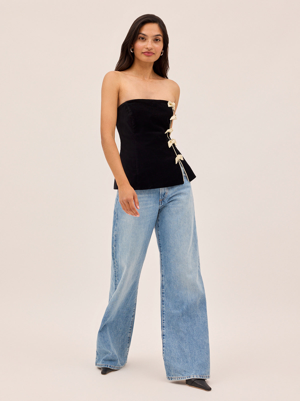 Bella Black Velvet Bandeau Top by Kitri Studio. A luxurious update on a KITRI favourite, this corset-inspired bandeau top is cut from black cotton velvet and reimagined for the winter party season.  Designed with striking ivory satin piping and a row of delicate satin bow details along one side, it offers both elegance and charm. strapless. Concealed side-zip. Fully lined. 100% Cotton Velvet.