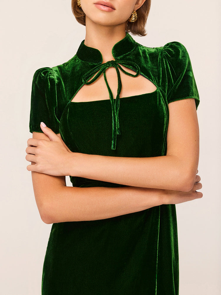 A person is dressed in the Carmella Green Velvet Midi Dress by KITRI, showcasing its forest green hue and elegant velvet texture. The dress boasts short puffed sleeves, a keyhole neckline accompanied by a tied bow at the neck, and a subtle Cheongsam-inspired design. Their arms are folded across their torso, with short hair and gold hoop earrings complementing against the plain light beige background.