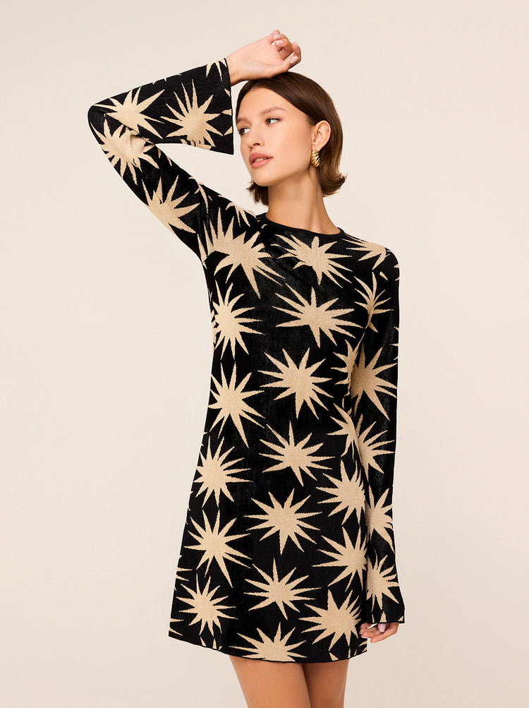 A woman stands against a neutral background, wearing the Greta Star Lurex Mini Knit Dress from KITRI. The black long-sleeve dress features metallic star prints that evoke a '70s vibe. She has short hair and accessorizes with gold hoop earrings, her right arm raised with her hand resting on her head as she gazes sideways. This outfit is a striking addition to any party wardrobe.